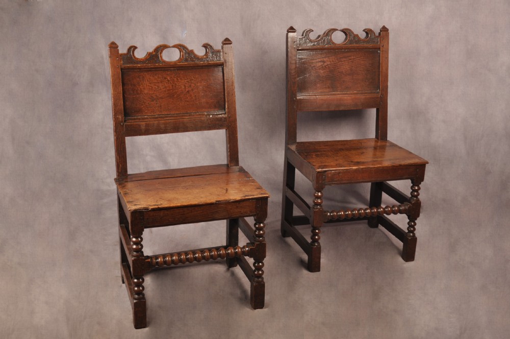 A pair of 17th century dark oak side chairs featuring carved crest rails, solid panel backs, and bobbin-turned front stretchers.