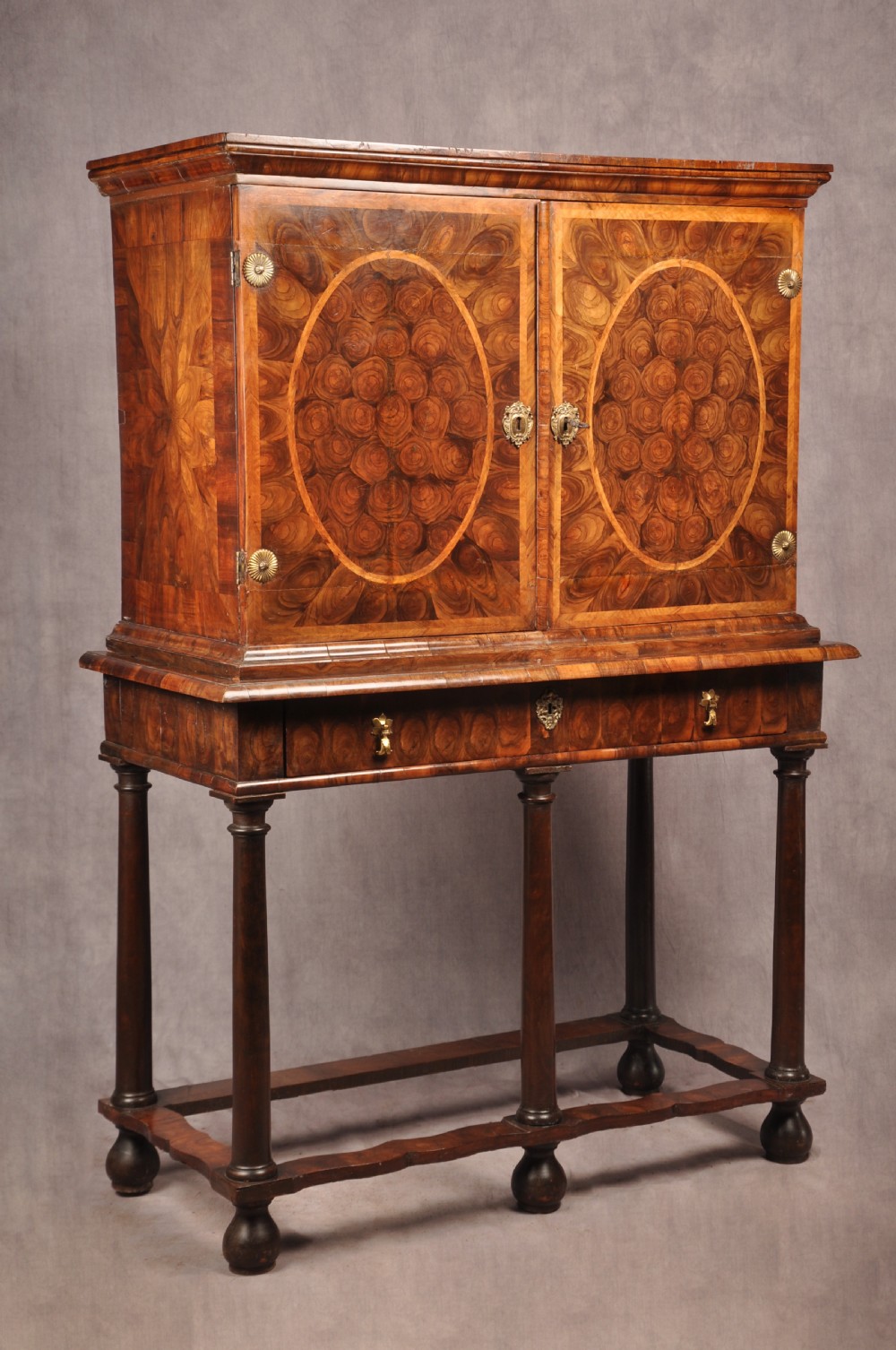 A 17th-century olive wood cabinet on stand featuring double doors inlaid with oval oyster veneer medallions, a lower drawer, turned legs, stretchers, and ball feet.