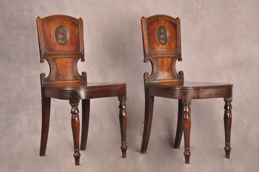 A pair of antique dark mahogany Regency hall chairs featuring solid seats, turned front legs, and shaped backrests decorated with oval armorial crest medallions.