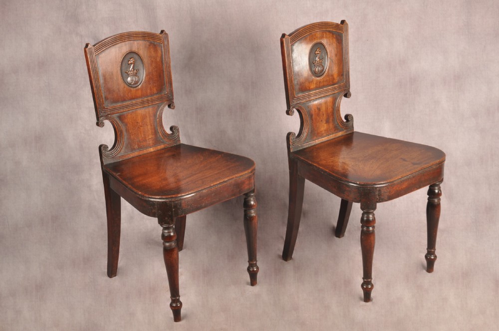 A pair of antique dark mahogany Regency hall chairs featuring solid seats, turned front legs, and shaped backrests decorated with oval armorial crest medallions. View II