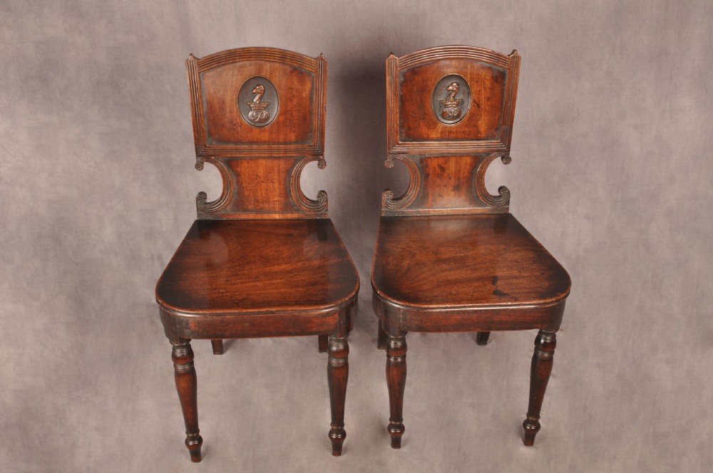 A pair of antique dark mahogany Regency hall chairs featuring solid seats, turned front legs, and shaped backrests decorated with oval armorial crest medallions. View III