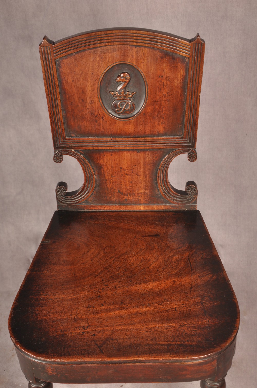 A pair of antique dark mahogany Regency hall chairs featuring solid seats, turned front legs, and shaped backrests decorated with oval armorial crest medallions. View IV