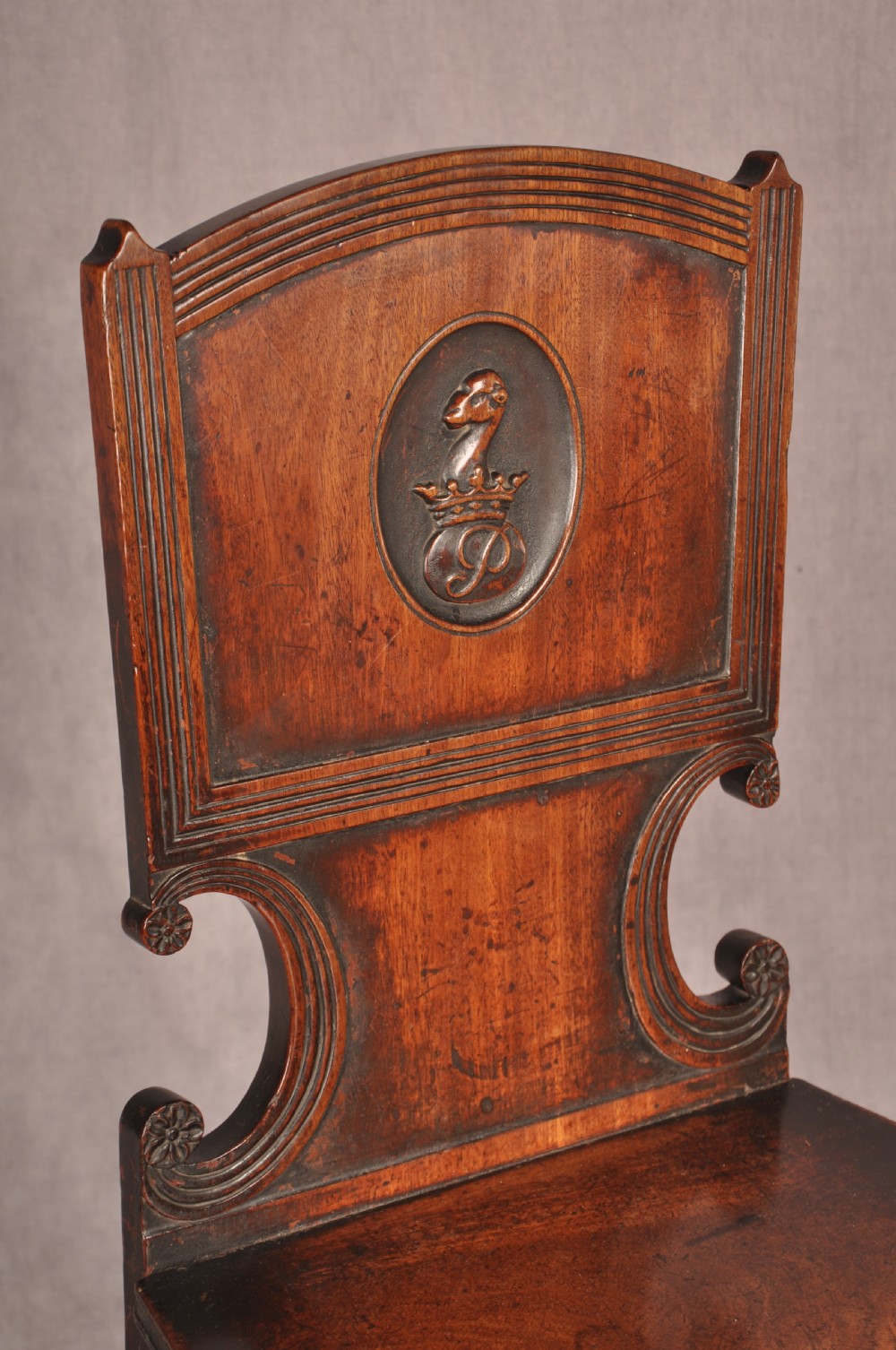 A pair of antique dark mahogany Regency hall chairs featuring solid seats, turned front legs, and shaped backrests decorated with oval armorial crest medallions. View V