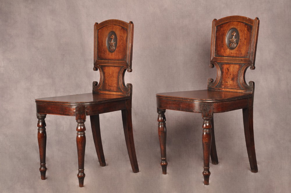A pair of antique dark mahogany Regency hall chairs featuring solid seats, turned front legs, and shaped backrests decorated with oval armorial crest medallions. View IX