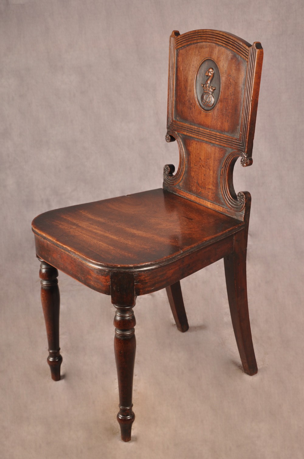 A pair of antique dark mahogany Regency hall chairs featuring solid seats, turned front legs, and shaped backrests decorated with oval armorial crest medallions. View X