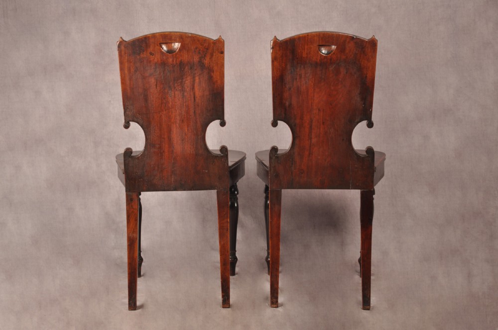 A pair of antique dark mahogany Regency hall chairs featuring solid seats, turned front legs, and shaped backrests decorated with oval armorial crest medallions. View XI