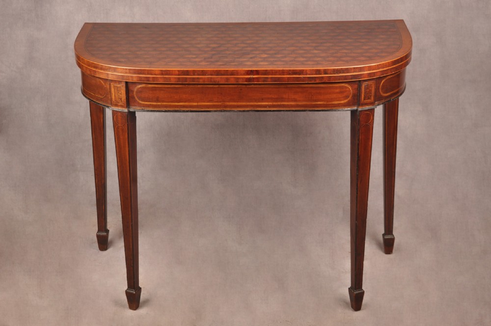 An 18th-century demilune card table featuring a geometric parquetry top, inlaid apron, and four square tapered legs ending in spade feet.