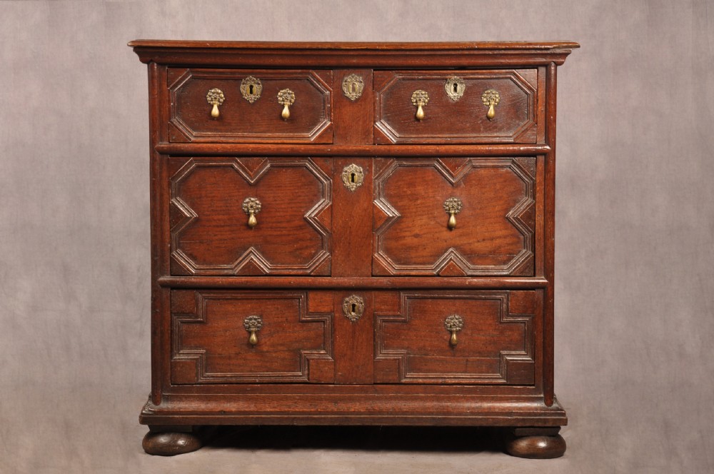 A Charles II dark oak chest of drawers featuring geometric moulded drawer panels, brass teardrop handles, keyhole escutcheons, and bun feet.
