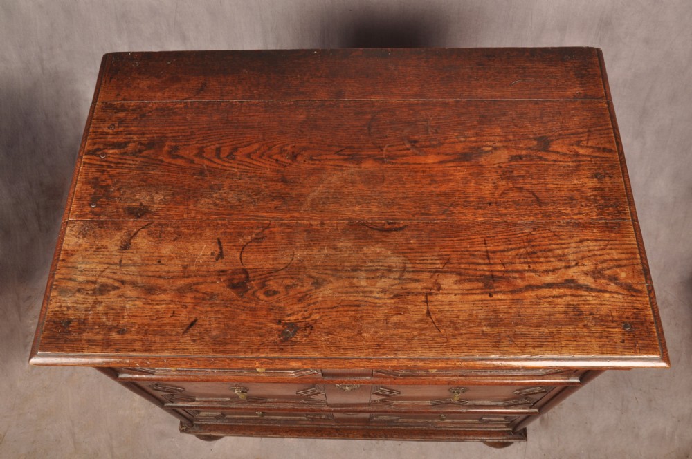 A Charles II dark oak chest of drawers featuring geometric moulded drawer panels, brass teardrop handles, keyhole escutcheons, and bun feet. View II