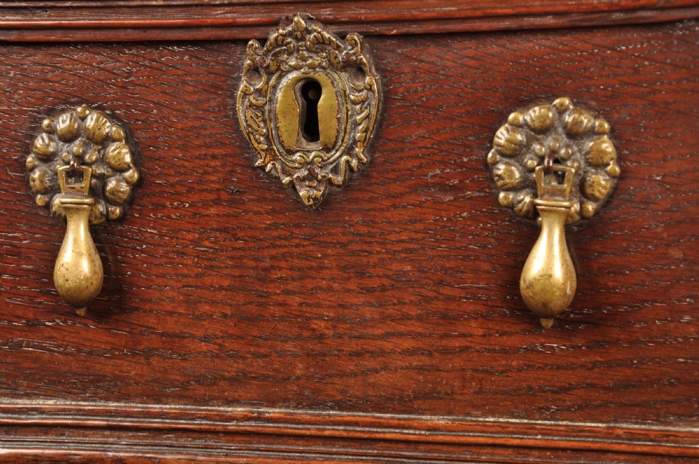 A Charles II dark oak chest of drawers featuring geometric moulded drawer panels, brass teardrop handles, keyhole escutcheons, and bun feet. View V