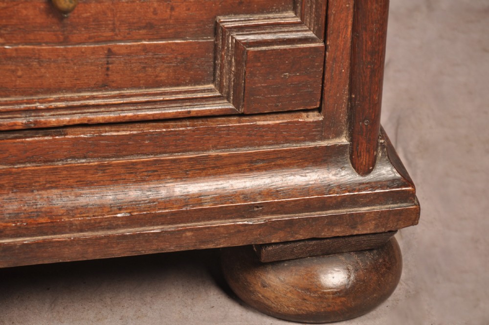 A Charles II dark oak chest of drawers featuring geometric moulded drawer panels, brass teardrop handles, keyhole escutcheons, and bun feet. View VII