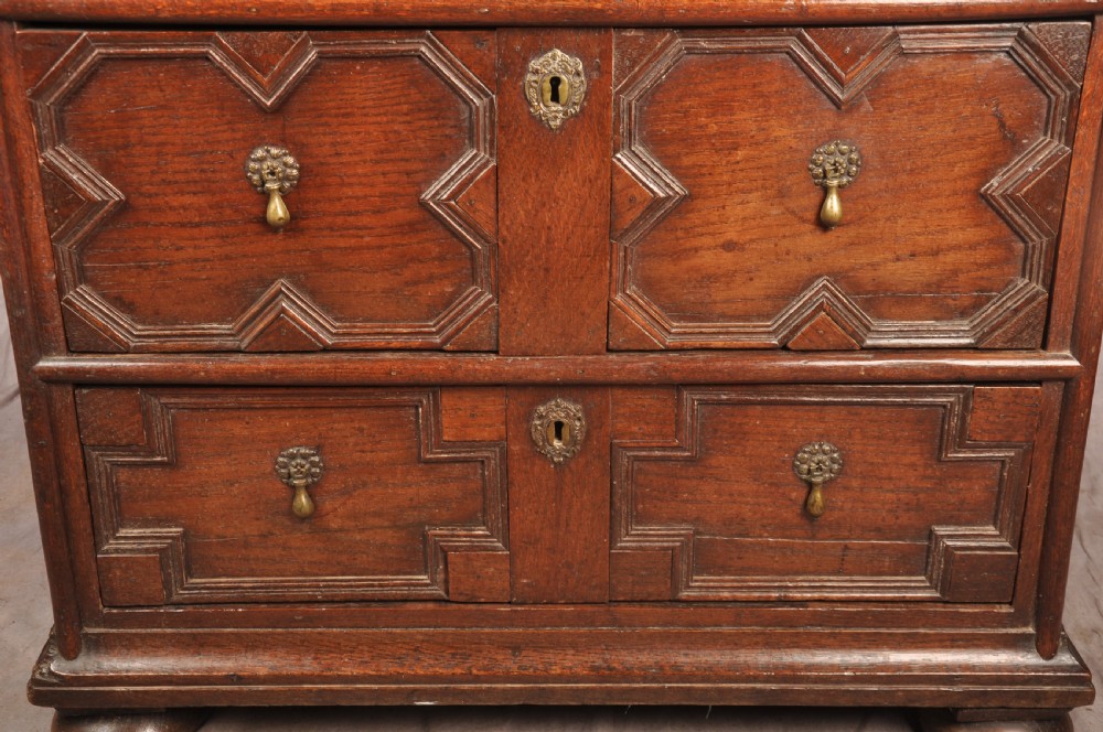 A Charles II dark oak chest of drawers featuring geometric moulded drawer panels, brass teardrop handles, keyhole escutcheons, and bun feet. View VIII