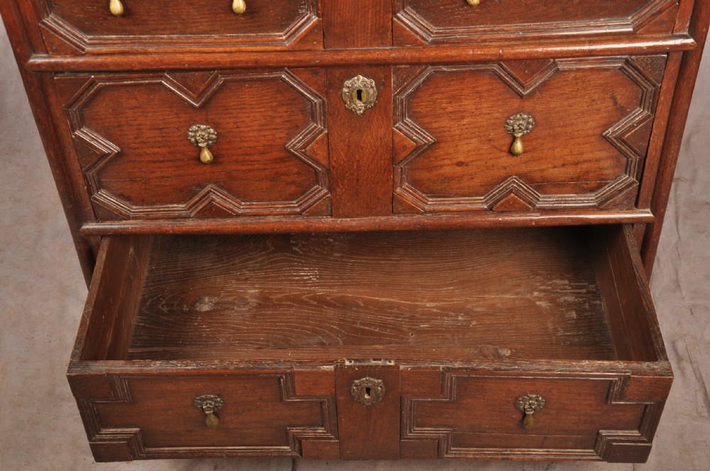 A Charles II dark oak chest of drawers featuring geometric moulded drawer panels, brass teardrop handles, keyhole escutcheons, and bun feet. View X