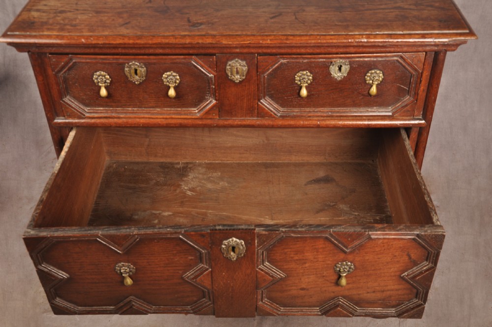 A Charles II dark oak chest of drawers featuring geometric moulded drawer panels, brass teardrop handles, keyhole escutcheons, and bun feet. View XI
