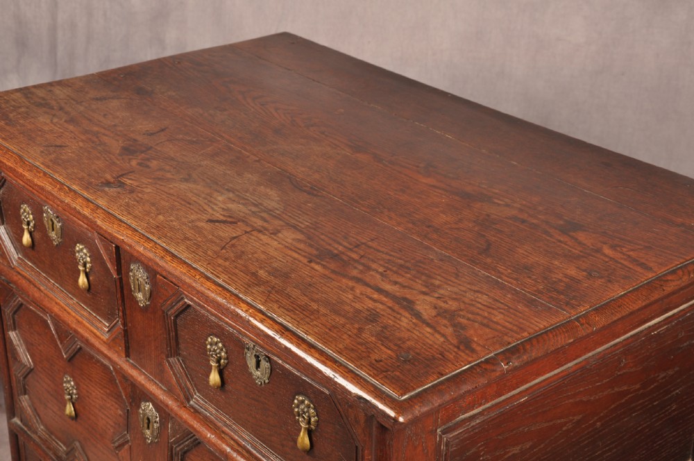 A Charles II dark oak chest of drawers featuring geometric moulded drawer panels, brass teardrop handles, keyhole escutcheons, and bun feet. View XVI