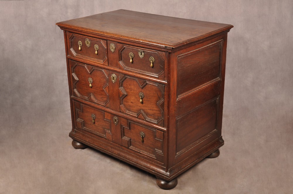 A Charles II dark oak chest of drawers featuring geometric moulded drawer panels, brass teardrop handles, keyhole escutcheons, and bun feet. View XIX