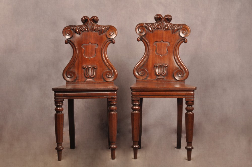 A pair of mahogany Regency hall chairs featuring carved scroll tops, shield motifs, flat timber seats, and turned front legs.
