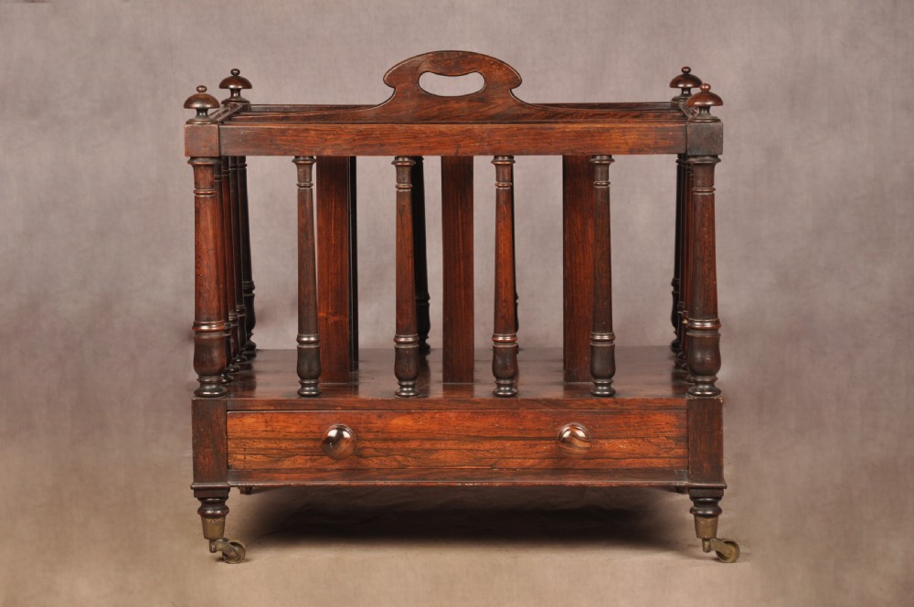 A Regency period rosewood Canterbury featuring four vertical slatted compartments, corner finials, a base drawer with wooden knobs, and brass castors.