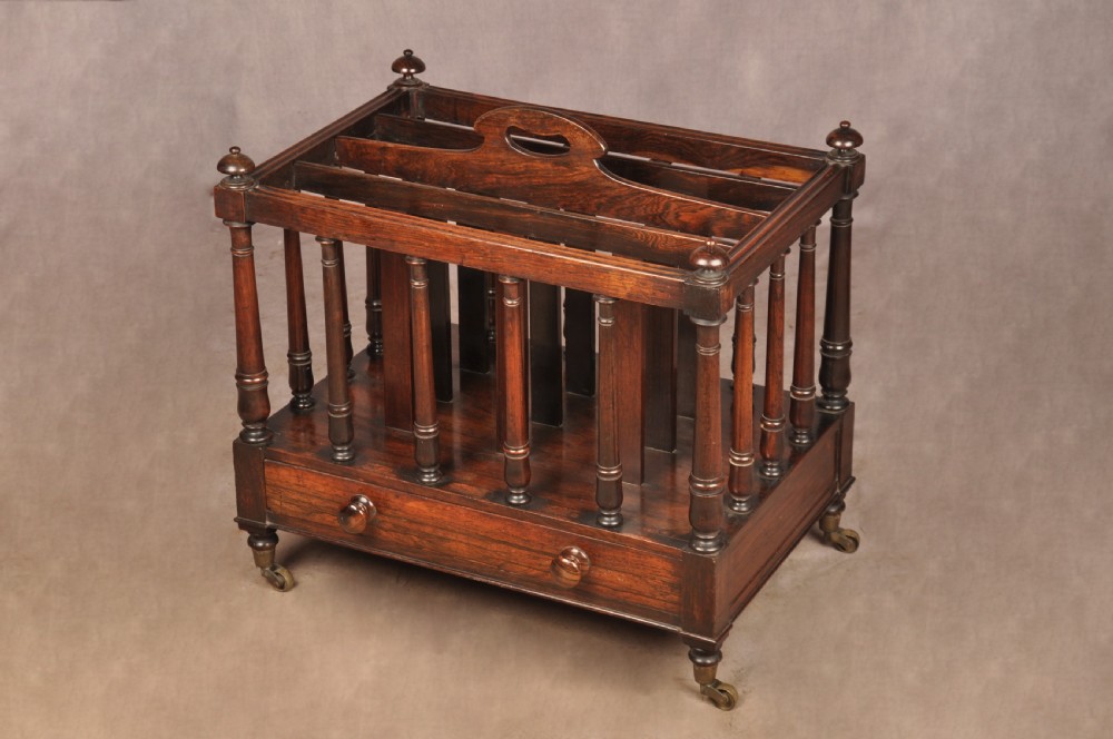 A Regency period rosewood Canterbury featuring four vertical slatted compartments, corner finials, a base drawer with wooden knobs, and brass castors. View IV