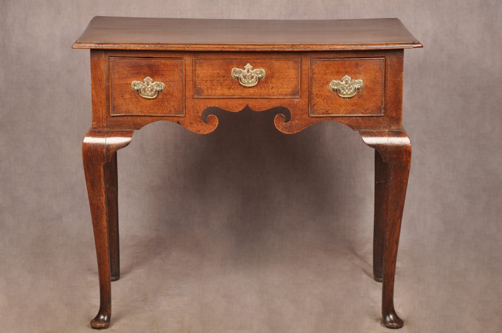 A mid-18th century George II oak lowboy side table featuring three drawers with brass handles, a shaped scroll apron, and cabriole legs ending in pad feet.