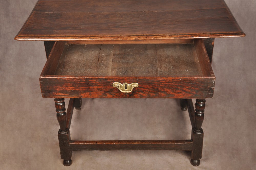 A 17th-century dark oak side table featuring a single drawer with a brass handle, turned legs, and a perimeter box stretcher. View X