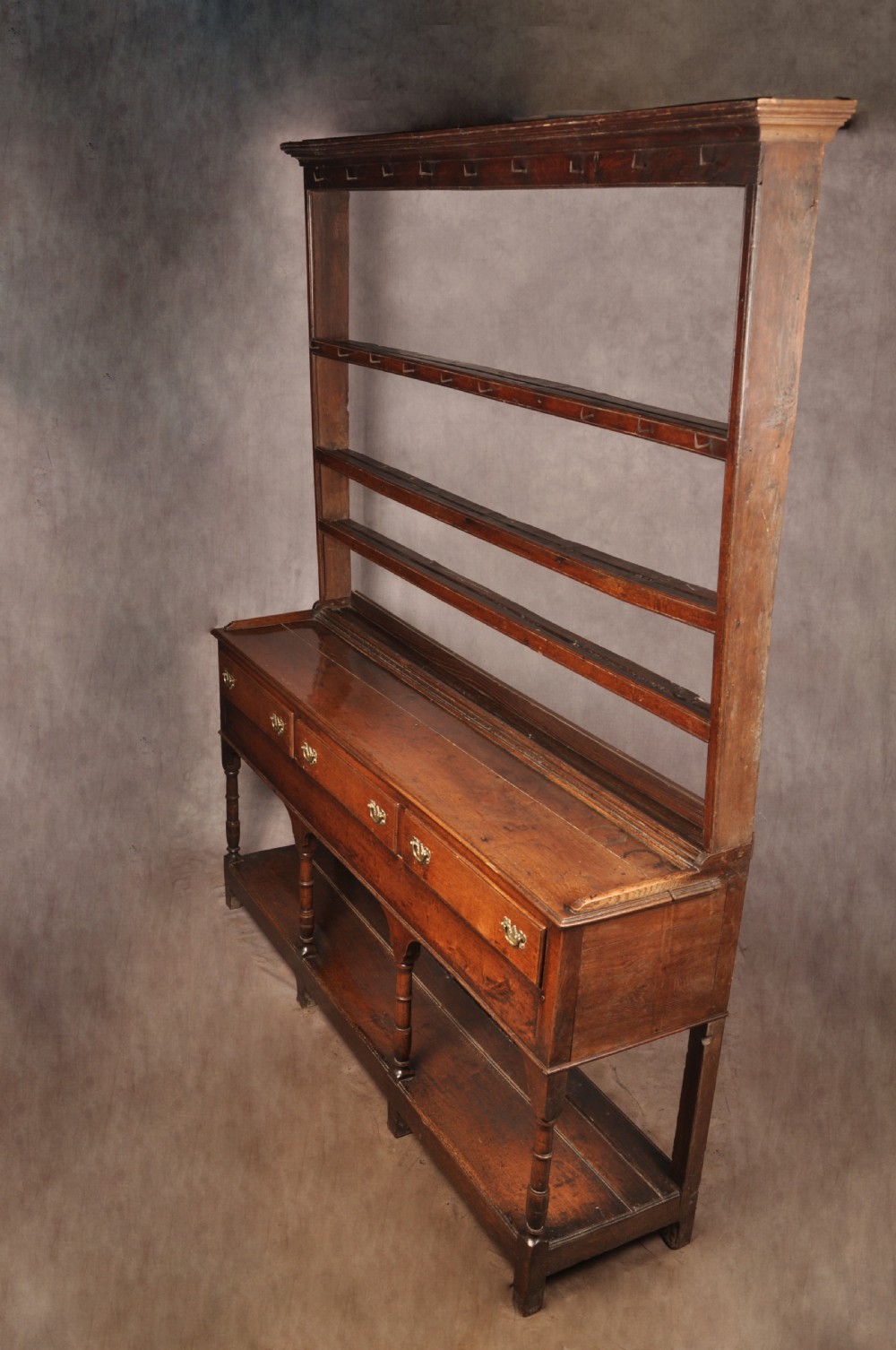 An 18th-century oak pot board dresser featuring an open tiered plate rack above three drawers with brass handles and a low shelf. View XVI