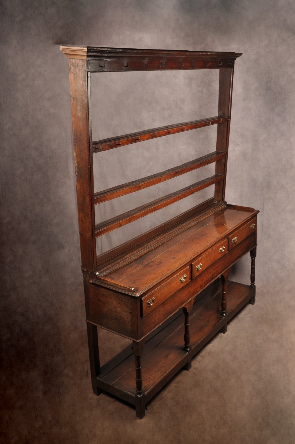 An 18th-century oak pot board dresser featuring an open tiered plate rack above three drawers with brass handles and a low shelf. View XVIII