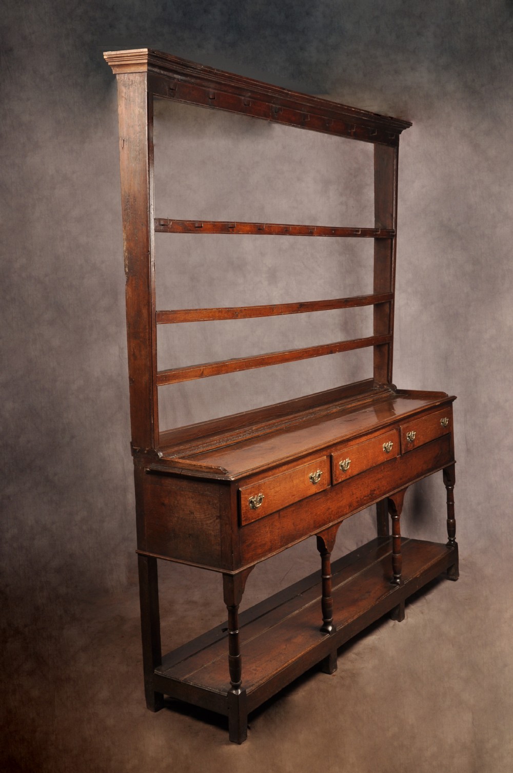 An 18th-century oak pot board dresser featuring an open tiered plate rack above three drawers with brass handles and a low shelf.