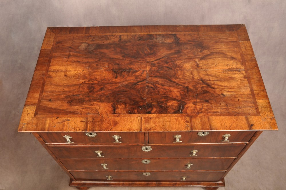 A 17th-century walnut chest of drawers featuring two short drawers over three long drawers, brass drop handles, keyhole escutcheons, and large turned bun feet. View V