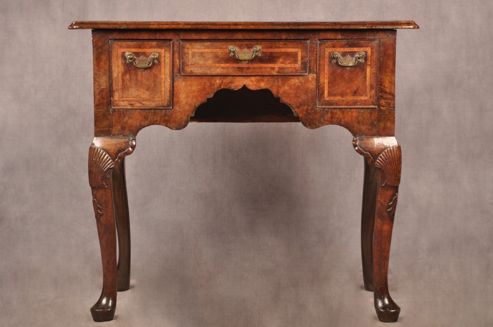 An early 18th century walnut lowboy featuring three drawers with brass handles, a shaped apron, and shell-carved cabriole legs.