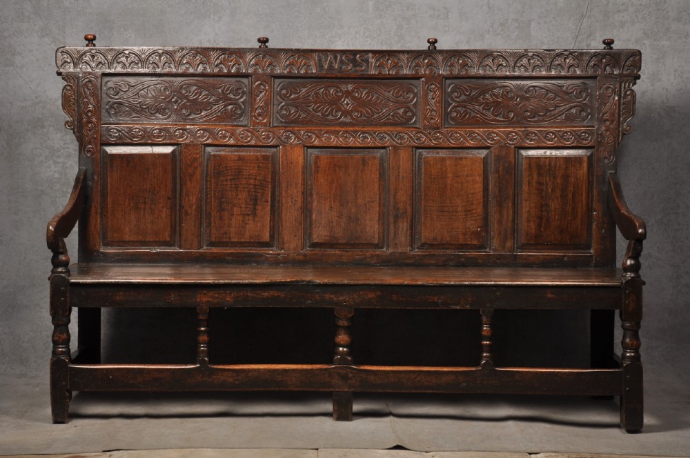 A 17th-century dark oak settle featuring a high carved panelled back with carved cresting and initials above a long bench seat with turned legs.