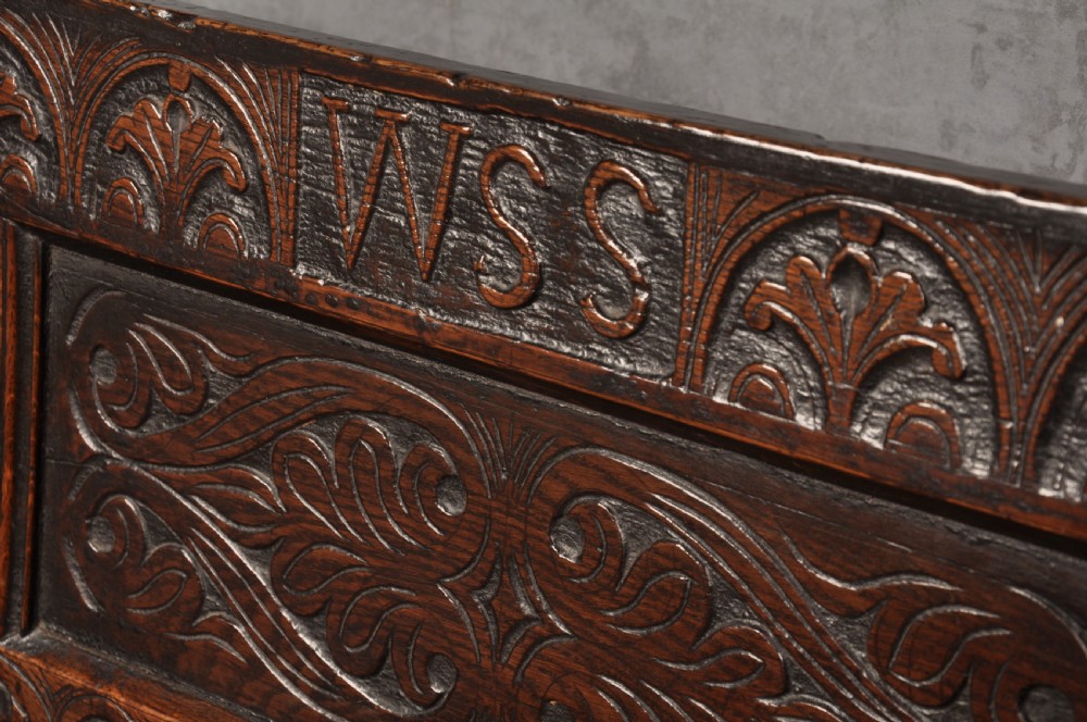A 17th-century dark oak settle featuring a high carved panelled back with carved cresting and initials above a long bench seat with turned legs. View II
