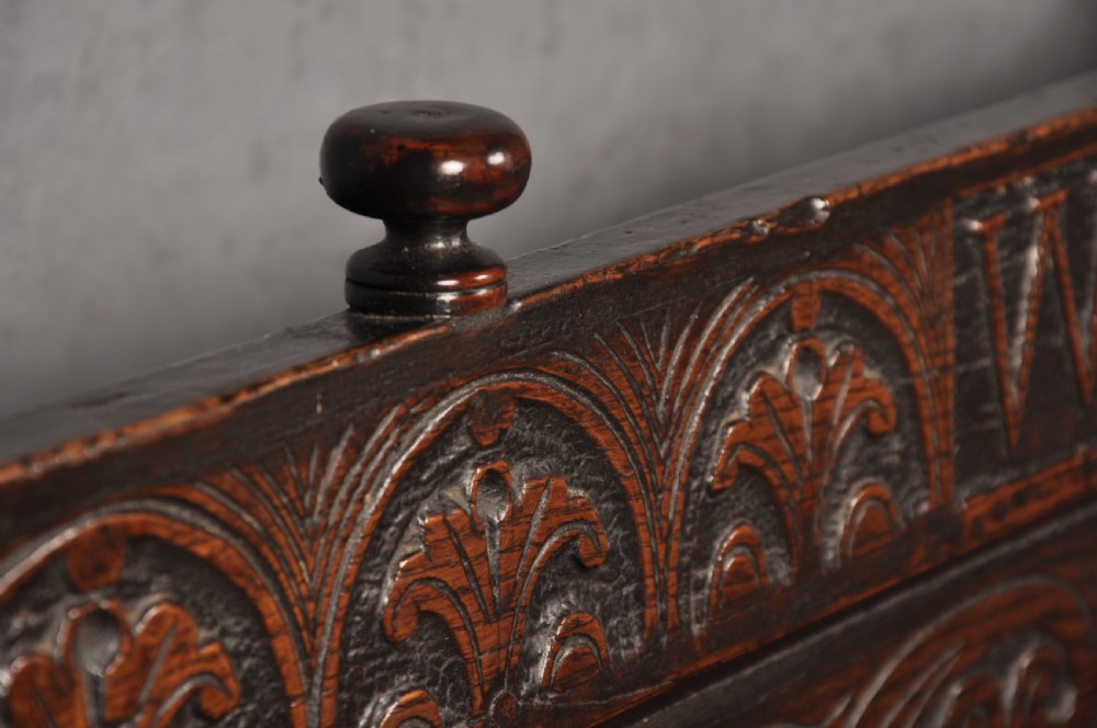 A 17th-century dark oak settle featuring a high carved panelled back with carved cresting and initials above a long bench seat with turned legs. View III