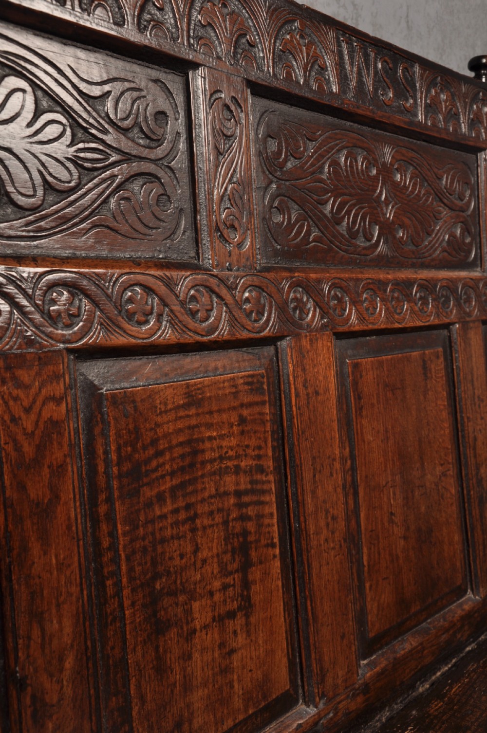 A 17th-century dark oak settle featuring a high carved panelled back with carved cresting and initials above a long bench seat with turned legs. View VI