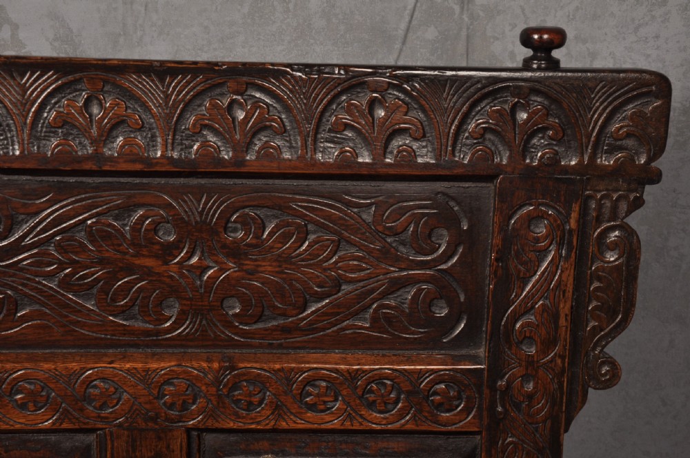 A 17th-century dark oak settle featuring a high carved panelled back with carved cresting and initials above a long bench seat with turned legs. View VII