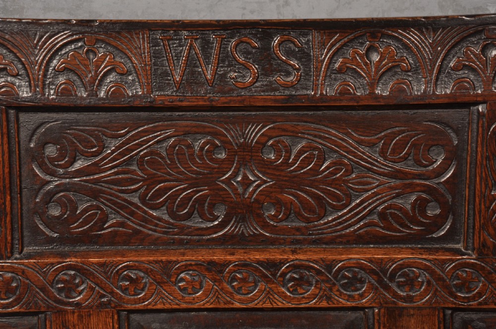 A 17th-century dark oak settle featuring a high carved panelled back with carved cresting and initials above a long bench seat with turned legs. View VIII