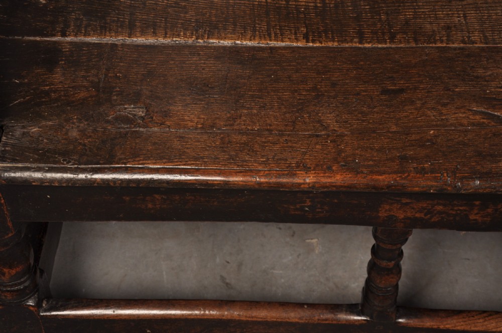 A 17th-century dark oak settle featuring a high carved panelled back with carved cresting and initials above a long bench seat with turned legs. View IX