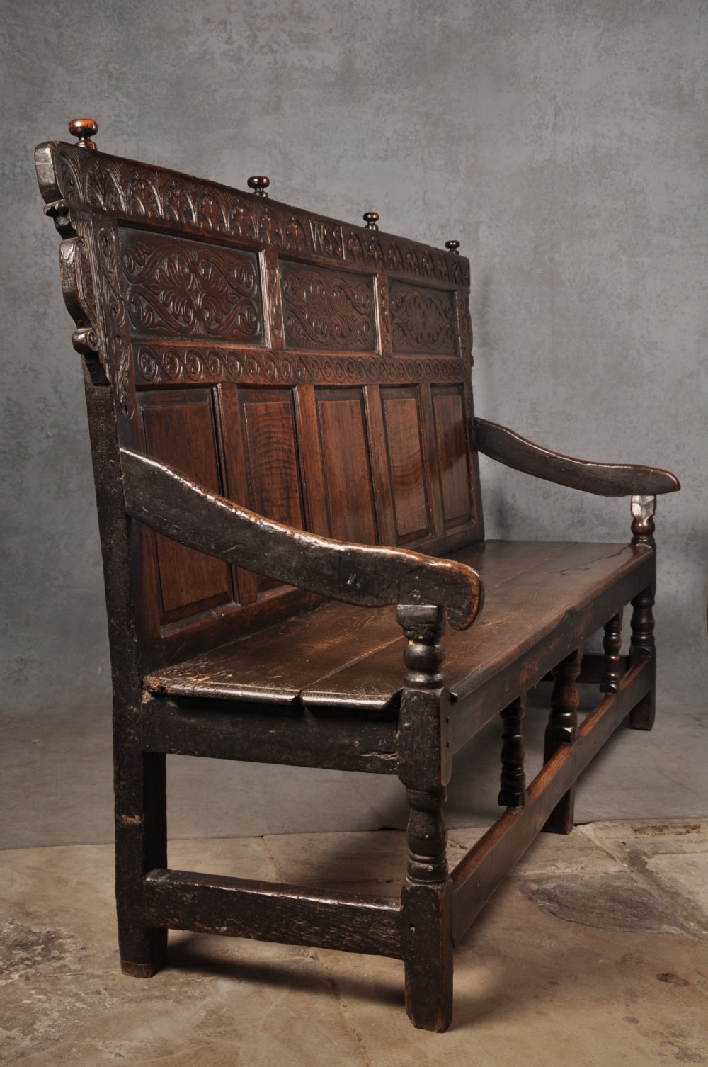 A 17th-century dark oak settle featuring a high carved panelled back with carved cresting and initials above a long bench seat with turned legs. View X