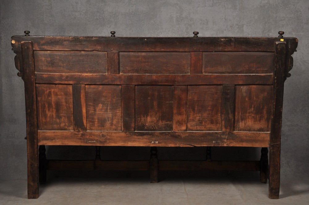 A 17th-century dark oak settle featuring a high carved panelled back with carved cresting and initials above a long bench seat with turned legs. View XI