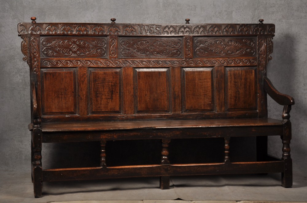 A 17th-century dark oak settle featuring a high carved panelled back with carved cresting and initials above a long bench seat with turned legs. View XV