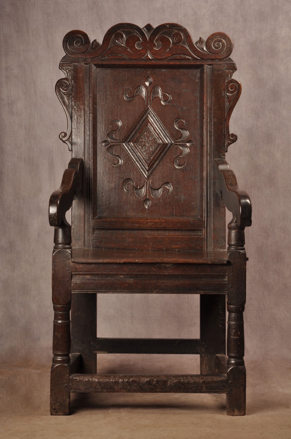 A 17th-century dark oak wainscot armchair featuring a carved scrolled cresting rail, a lozenge-carved back panel, turned front legs, and a solid wooden seat.
