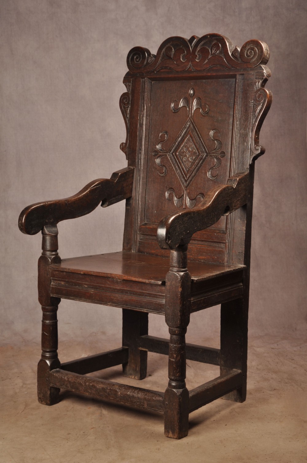 A 17th-century dark oak wainscot armchair featuring a carved scrolled cresting rail, a lozenge-carved back panel, turned front legs, and a solid wooden seat. View IV