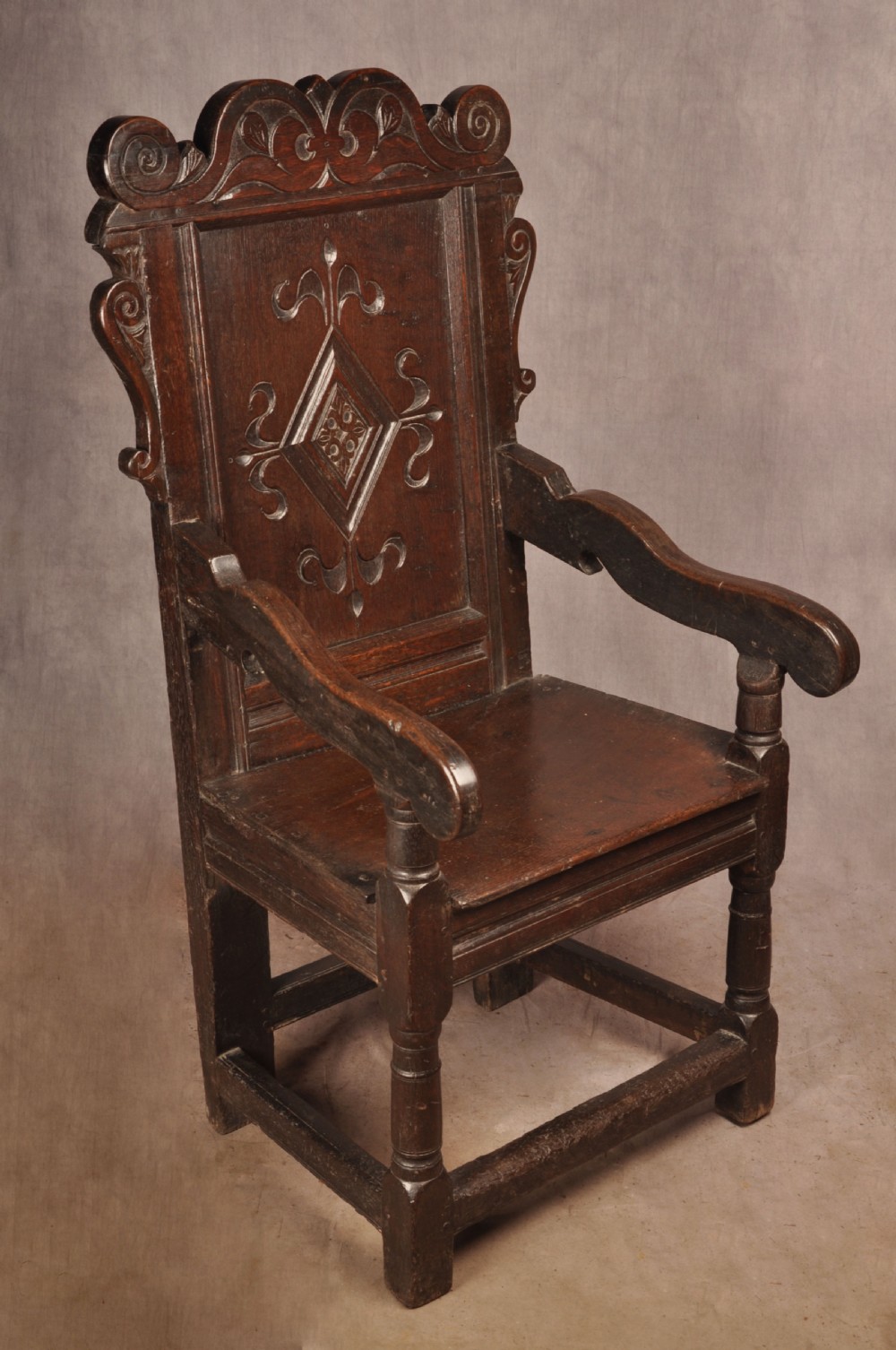 A 17th-century dark oak wainscot armchair featuring a carved scrolled cresting rail, a lozenge-carved back panel, turned front legs, and a solid wooden seat. View V