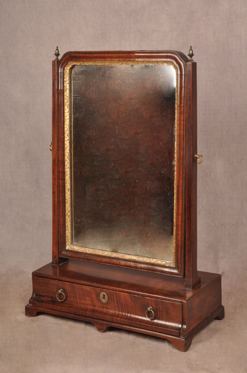 An 18th-century mahogany toilet mirror featuring an adjustable glass plate surrounded by a gilt slip, supported by vertical uprights with brass finials over a single-drawer plinth base.