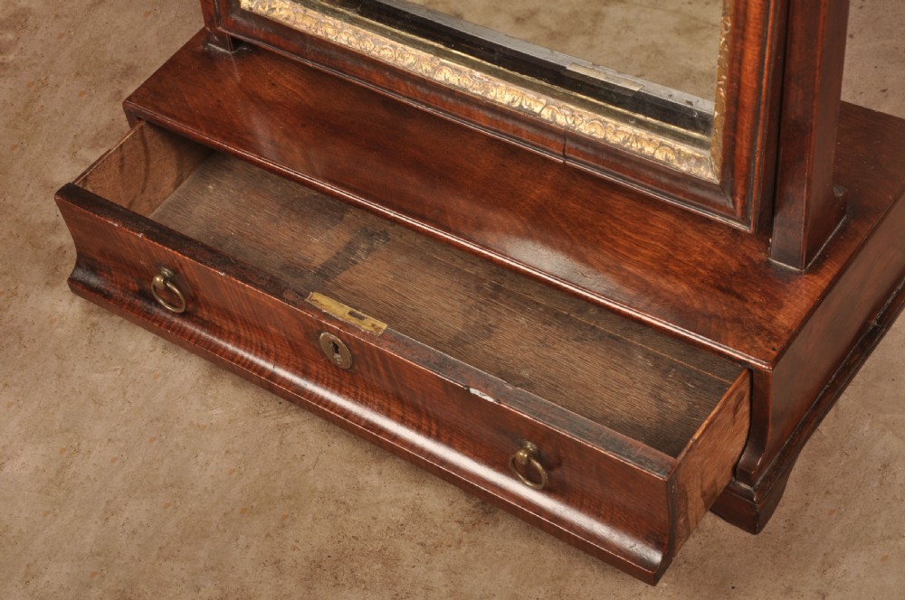 An 18th-century mahogany toilet mirror featuring an adjustable glass plate surrounded by a gilt slip, supported by vertical uprights with brass finials over a single-drawer plinth base. View IV