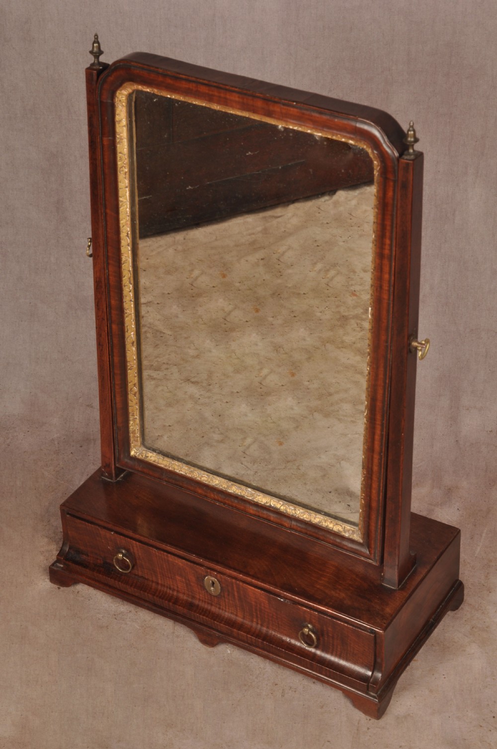 An 18th-century mahogany toilet mirror featuring an adjustable glass plate surrounded by a gilt slip, supported by vertical uprights with brass finials over a single-drawer plinth base. View VI