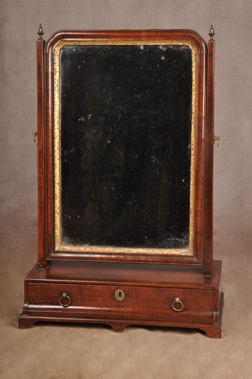 An 18th-century mahogany toilet mirror featuring an adjustable glass plate surrounded by a gilt slip, supported by vertical uprights with brass finials over a single-drawer plinth base. View VII
