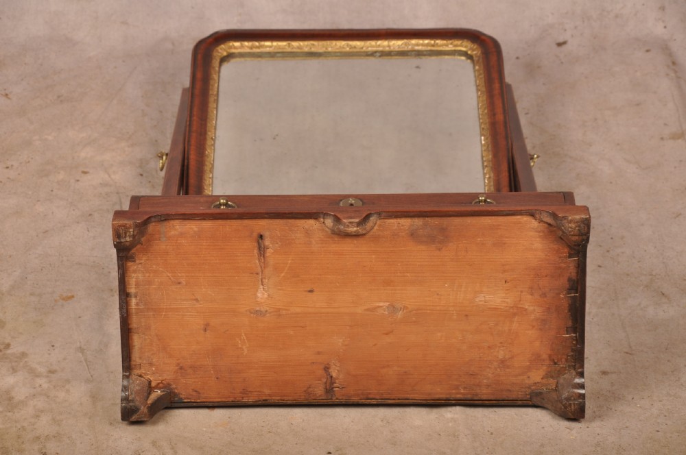 An 18th-century mahogany toilet mirror featuring an adjustable glass plate surrounded by a gilt slip, supported by vertical uprights with brass finials over a single-drawer plinth base. View IX