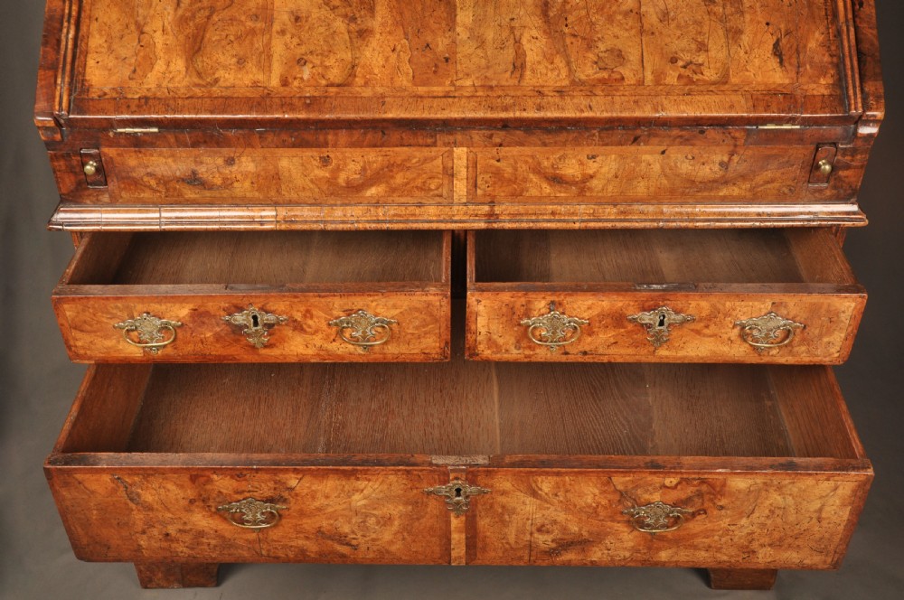 An eighteenth-century Queen Anne burr walnut bureau with a sloping fall-front lid above drawers featuring brass drop handles, resting on bracket feet. View VI