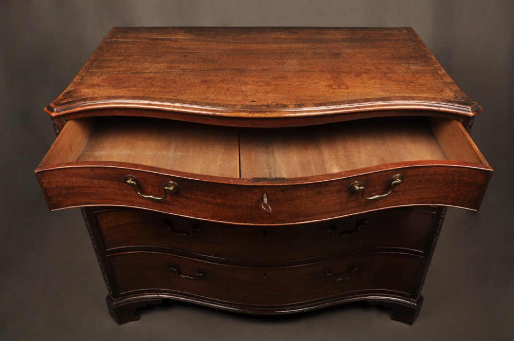 An 18th-century mahogany serpentine chest of drawers featuring four graduated drawers, carved canted corners, and brass swan-neck handles. View IX
